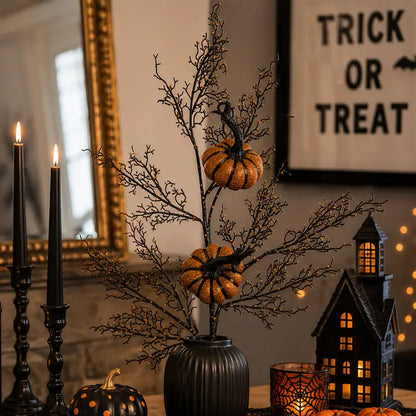 Halloween decorative centerpiece with black branch-like stems, two small orange pumpkins, and a ribbed black vase, styled on a tabletop with candles, a haunted house lantern, and a framed 'Trick or Treat' sign in the background.