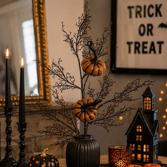 Halloween decorative centerpiece with black branch-like stems, two small orange pumpkins, and a ribbed black vase, styled on a tabletop with candles, a haunted house lantern, and a framed 'Trick or Treat' sign in the background.