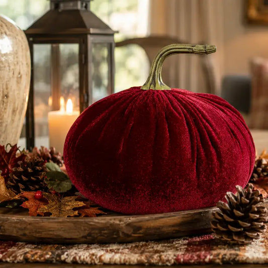 A plush red velvet decorative pumpkin with a faux stem, displayed on a rustic wooden tray with pinecones and autumn leaves, with a blurred candle lantern in the background.