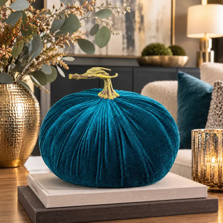 A teal velvet decorative pumpkin with a gold stem sits on stacked books in a styled living room setting.