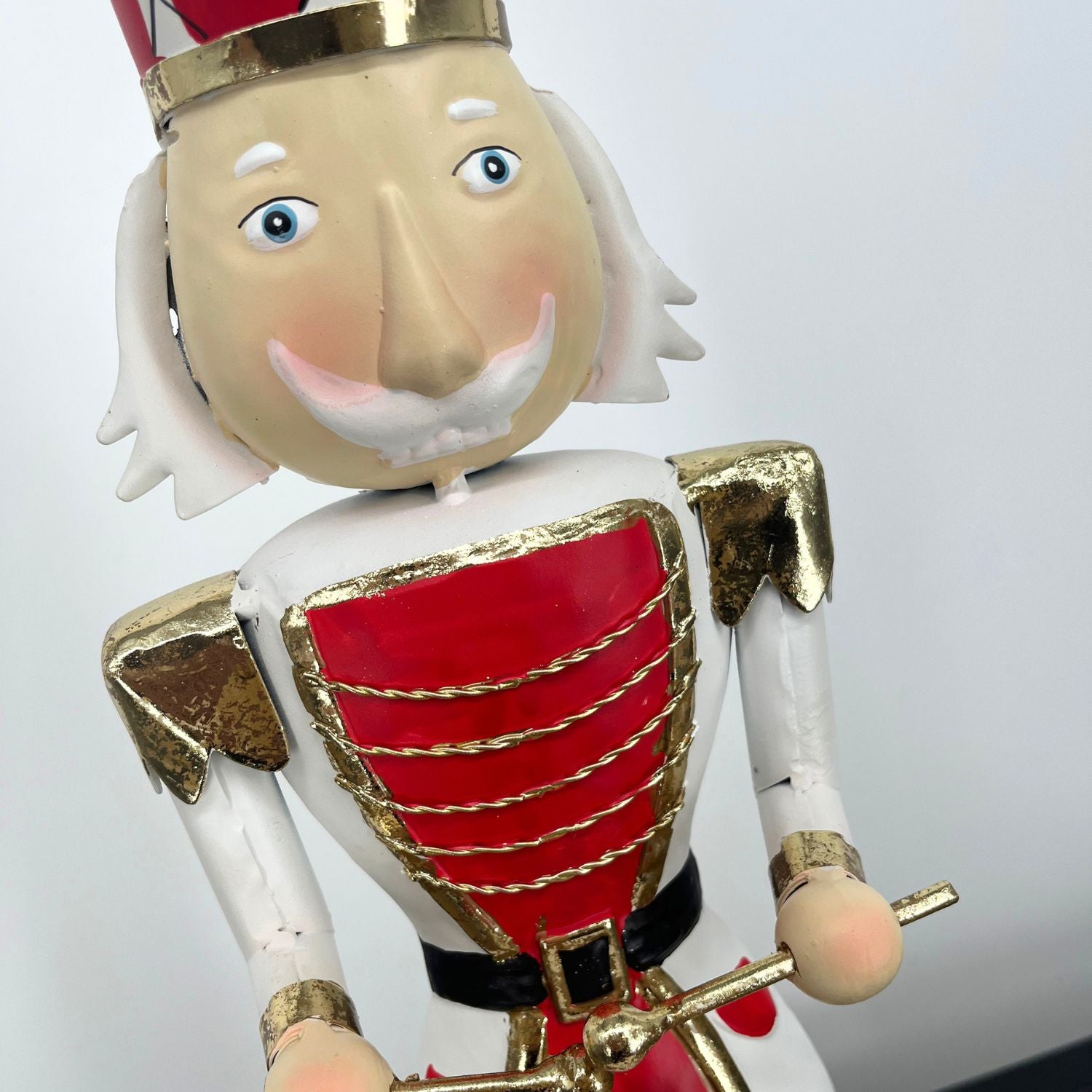 A traditional metal Christmas nutcracker figurine dressed in a red and white uniform with gold trim, a red and white striped hat, and black boots, holding a small drum against a plain white background.