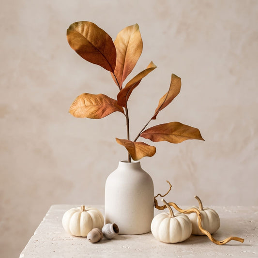 Autumn artificial magnolia foliage with large tan and brown leaves in a small matte white ceramic vase, styled on a stone surface with small white pumpkins and acorns against a warm beige background.