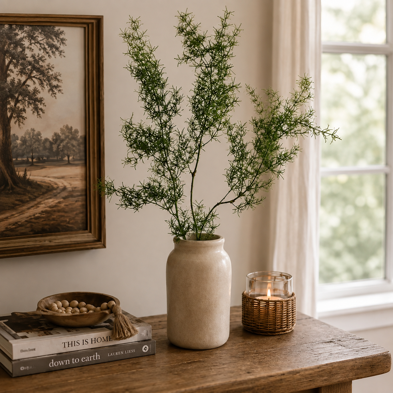 A tall beige ceramic vase holding delicate green foliage branches, styled on a rustic wooden table beside a lit woven candle holder and stacked books, with a framed landscape painting and window in the background.