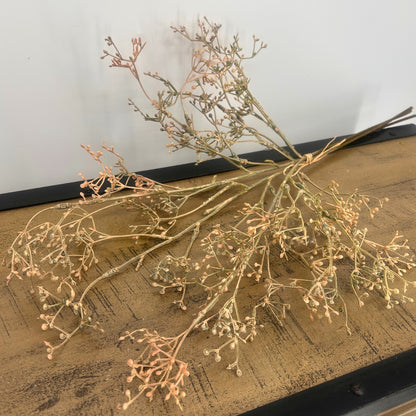 A bundle of dried baby's breath flowers with delicate pale pink and beige stems, arranged on a rustic wooden surface against a plain light gray wall.