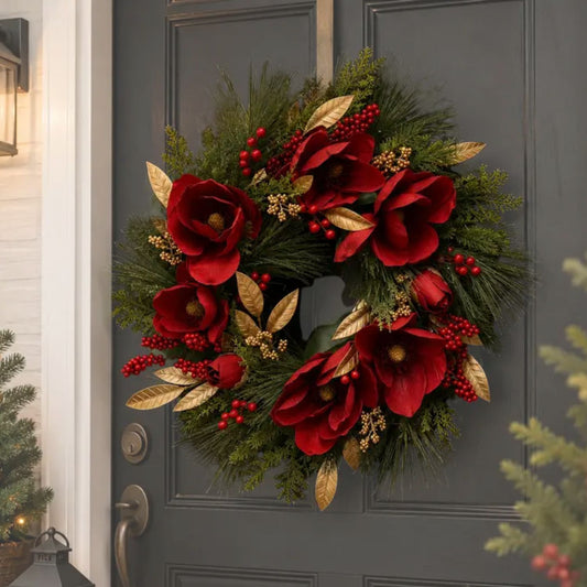 A festive artificial Christmas wreath hanging on a dark gray front door. The wreath features large red magnolia flowers, green pine branches, red berry clusters, and metallic gold leaves.