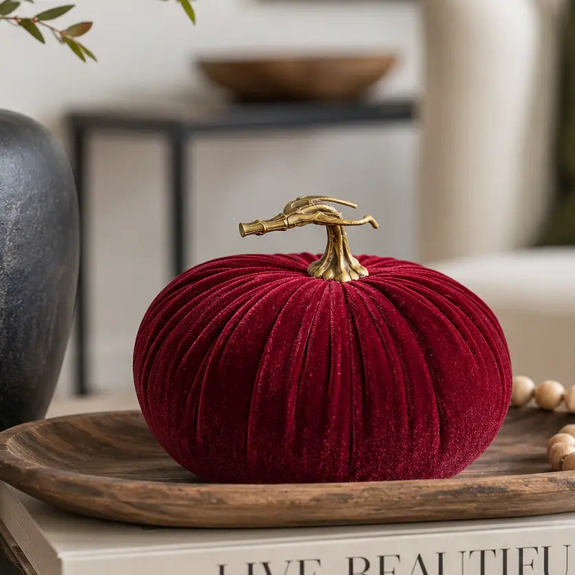 A deep red velvet decorative pumpkin with a realistic stem, displayed on a wooden tray with books and beads in a styled home setting.
