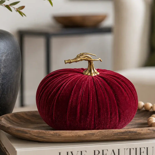 A deep red velvet decorative pumpkin with a realistic stem, displayed on a wooden tray with books and beads in a styled home setting.