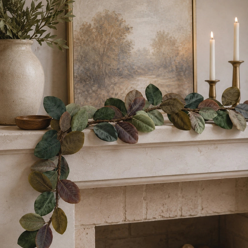 A faux magnolia leaf garland with realistic variegated leaves draped across a cream-colored fireplace mantel, styled with a ceramic vase, a framed landscape painting, and lit candles.