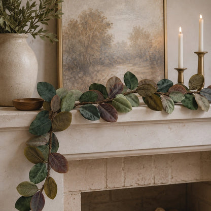 A faux magnolia leaf garland with realistic variegated leaves draped across a cream-colored fireplace mantel, styled with a ceramic vase, a framed landscape painting, and lit candles.