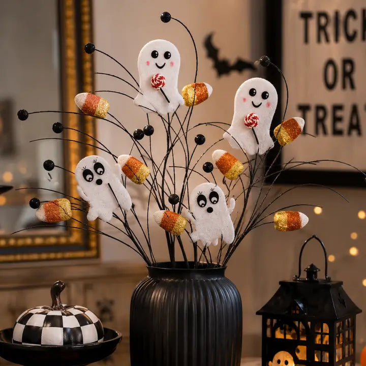 Halloween decorative stems with three white ghost cutouts holding candy lollipops, several orange-and-yellow candy corn pieces, and black glitter wire stems arranged in a ribbed black vase. The scene is styled indoors with warm lighting, a pumpkin, lantern, and blurred sign in the background.