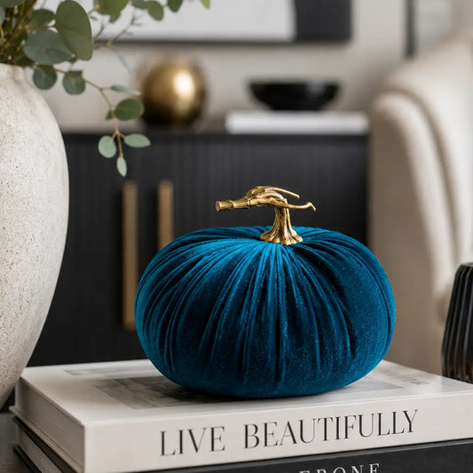 A teal velvet decorative pumpkin with a realistic stem sits on stacked books in a styled home interior setting.
