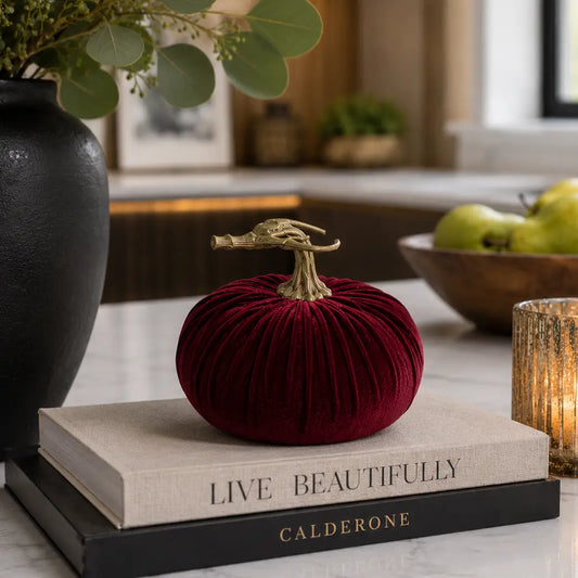 A small velvet decorative pumpkin in deep burgundy with a realistic stem, styled on top of two books on a kitchen counter.
