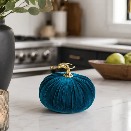 A small teal velvet decorative pumpkin with a polished gold metal stem, styled on a white marble kitchen countertop.